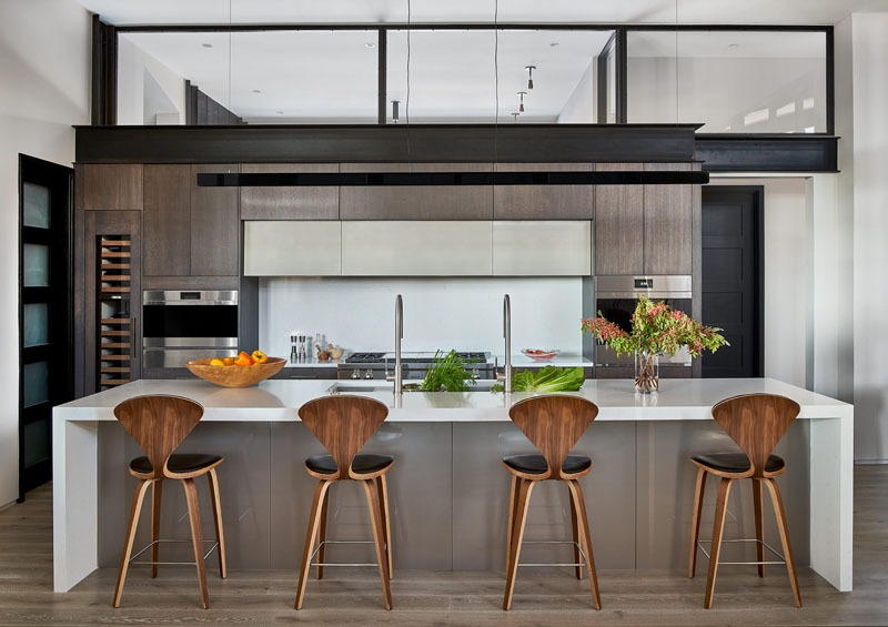 Modern kitchen with a central island and bar stools, dark wood cabinetry, stainless steel appliances, and an overhead metal frame. fresh fruit and flowers add color to the neutral palette.