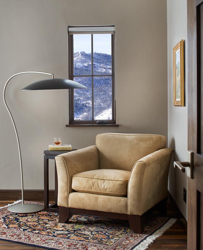 A cozy room with a tan armchair beside a small round table with a drink, against a backdrop of a window showing a snowy mountain landscape. a floor lamp curves overhead and an ornate rug covers the floor.