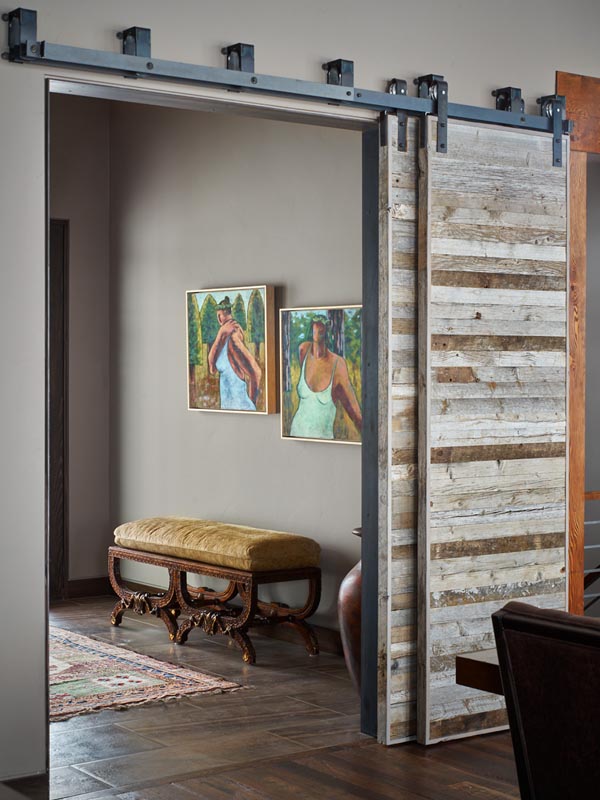 A rustic sliding barn door, partially open, revealing a small corridor with two framed paintings of women, and a vintage bench set against a decorative rug.