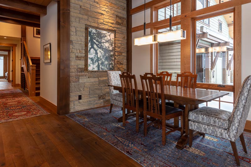 A cozy, well-lit dining area featuring a wooden table with six chairs, set on a vibrant rug, adjacent to large, bright windows in a room accented with stone walls and wooden beams.