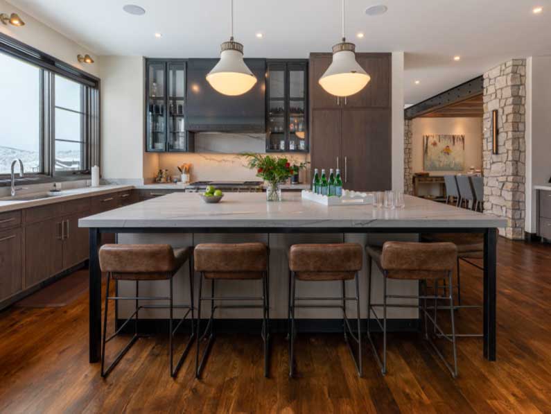 Modern kitchen with a large central island featuring a marble countertop and three brown bar stools. dark wood cabinets, white walls, a stone column, and hanging pendant lights complete the scene.