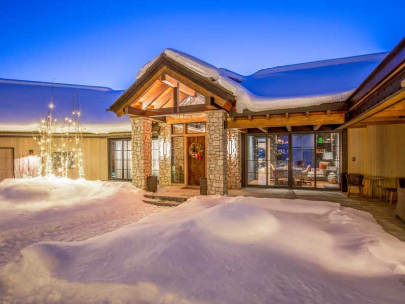 A cozy snow-covered house illuminated at twilight, featuring a stone facade and warm lighting from the windows, set in a serene winter landscape.