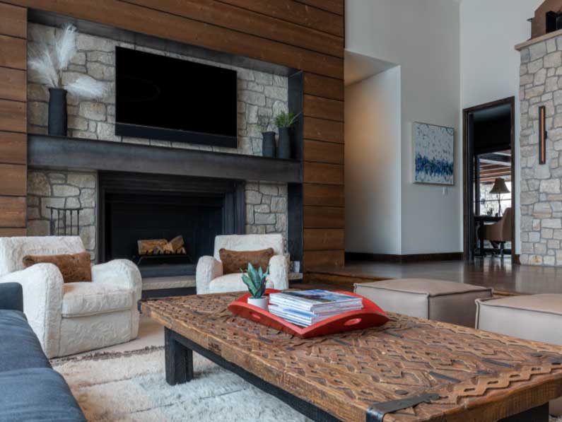 Cozy living room with a stone fireplace, wooden walls, a plush sofa, and armchairs. a patterned wooden coffee table with books and a plant is in the foreground.