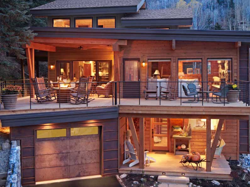 A cozy, multi-level wooden home illuminated at twilight, featuring a spacious deck with seating and visible warm interiors through large windows. the house is surrounded by a forested area.