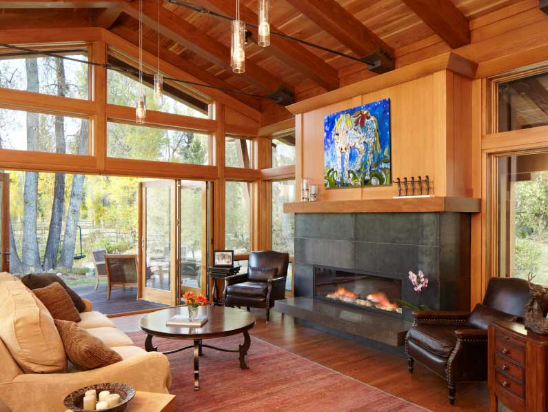 A cozy living room featuring a large stone fireplace, wooden beams, and floor-to-ceiling windows overlooking a forest. furnished with a mix of modern and traditional elements.