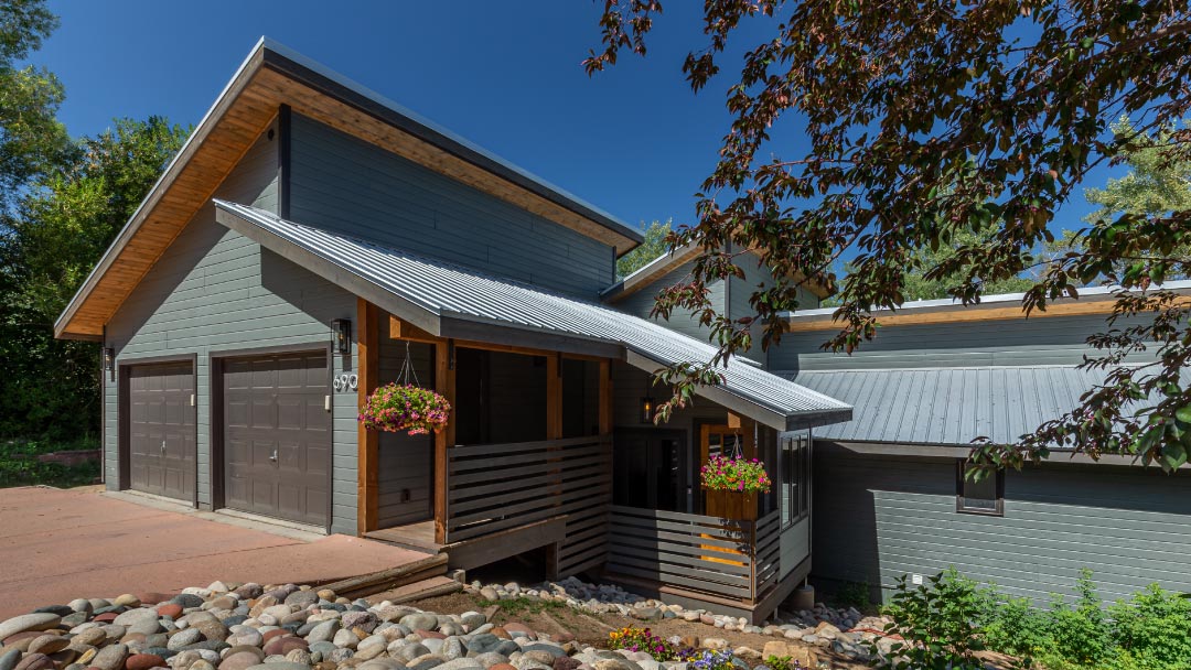 A contemporary home with a gray façade, metal roofing, and attached garage, surrounded by lush greenery and adorned with hanging flower baskets.