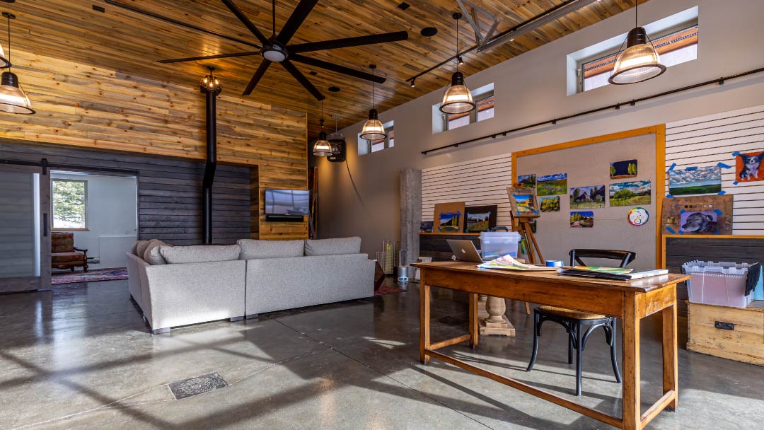 A spacious modern office with a wooden desk, a gray sofa, colorful wall art, and ceiling fans, featuring wooden beams and concrete flooring.