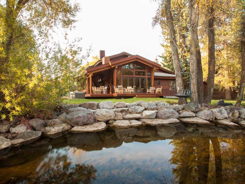 A serene view of a rustic wooden cabin with an expansive porch, surrounded by trees, reflecting in the tranquil water of a small pond bordered by a stone wall.